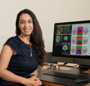 A smiling woman with long dark hair, wearing a black short-sleeved top is seated at a desk with a computer. Colorful images of brain structures are on the computer screen.